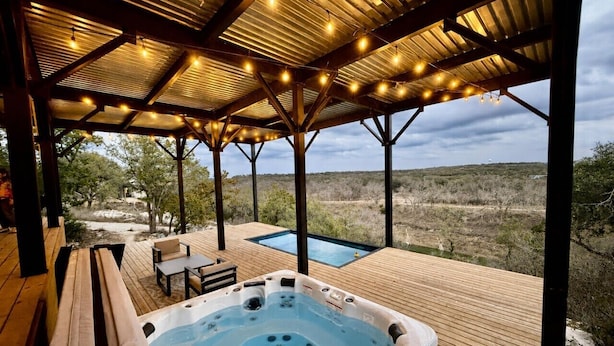 Een hottub op een houten terras, met een zwembad, omringd door een overdekt gebied met lichtjes.