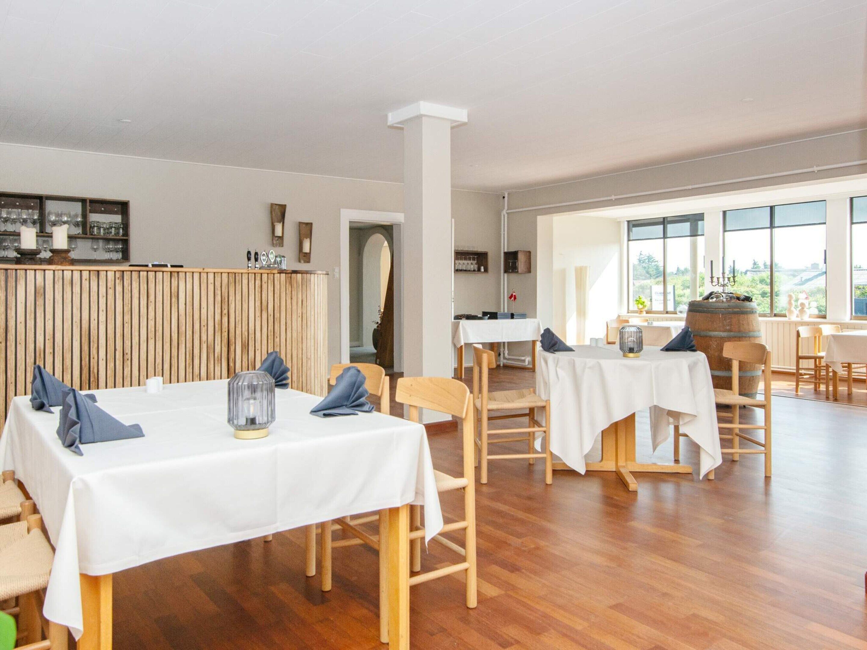 A spacious dining area with wooden flooring, white tablecloths, and simple wooden chairs.
