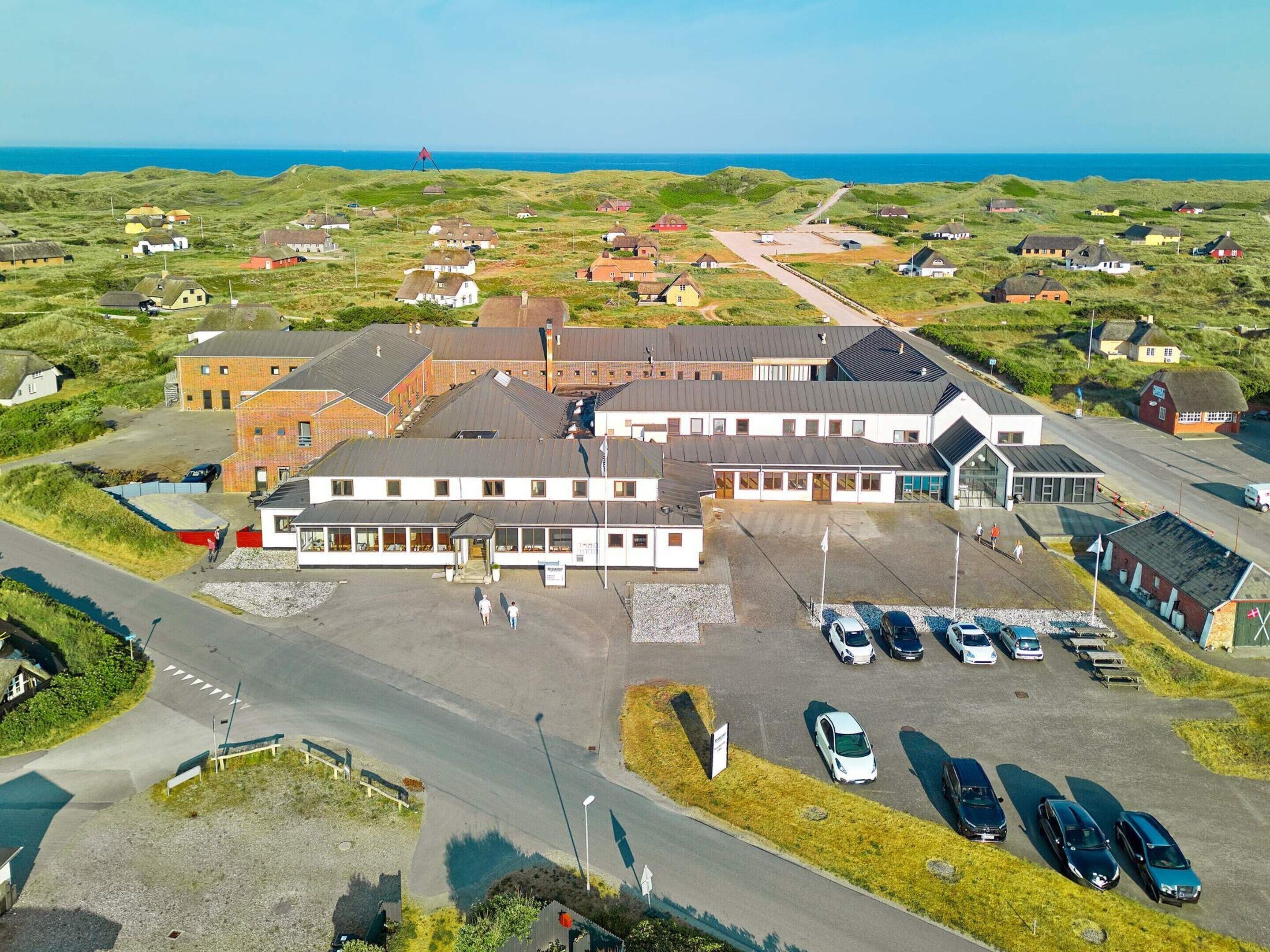 An aerial view of a building complex with a parking lot, surrounded by grassy fields and houses.