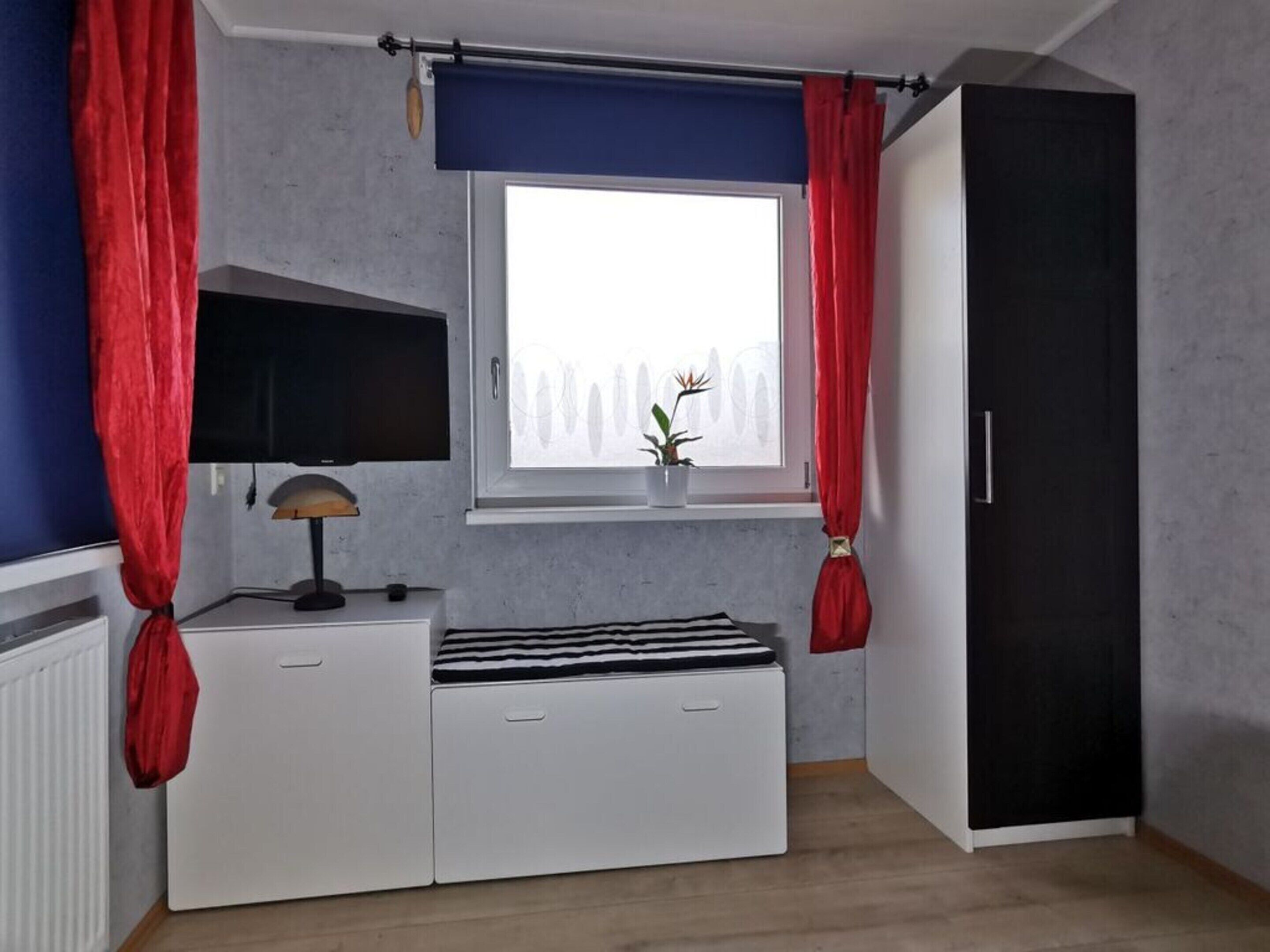 A room with a bed, a window with curtains, a radiator, a desk with a lamp, and a wardrobe.