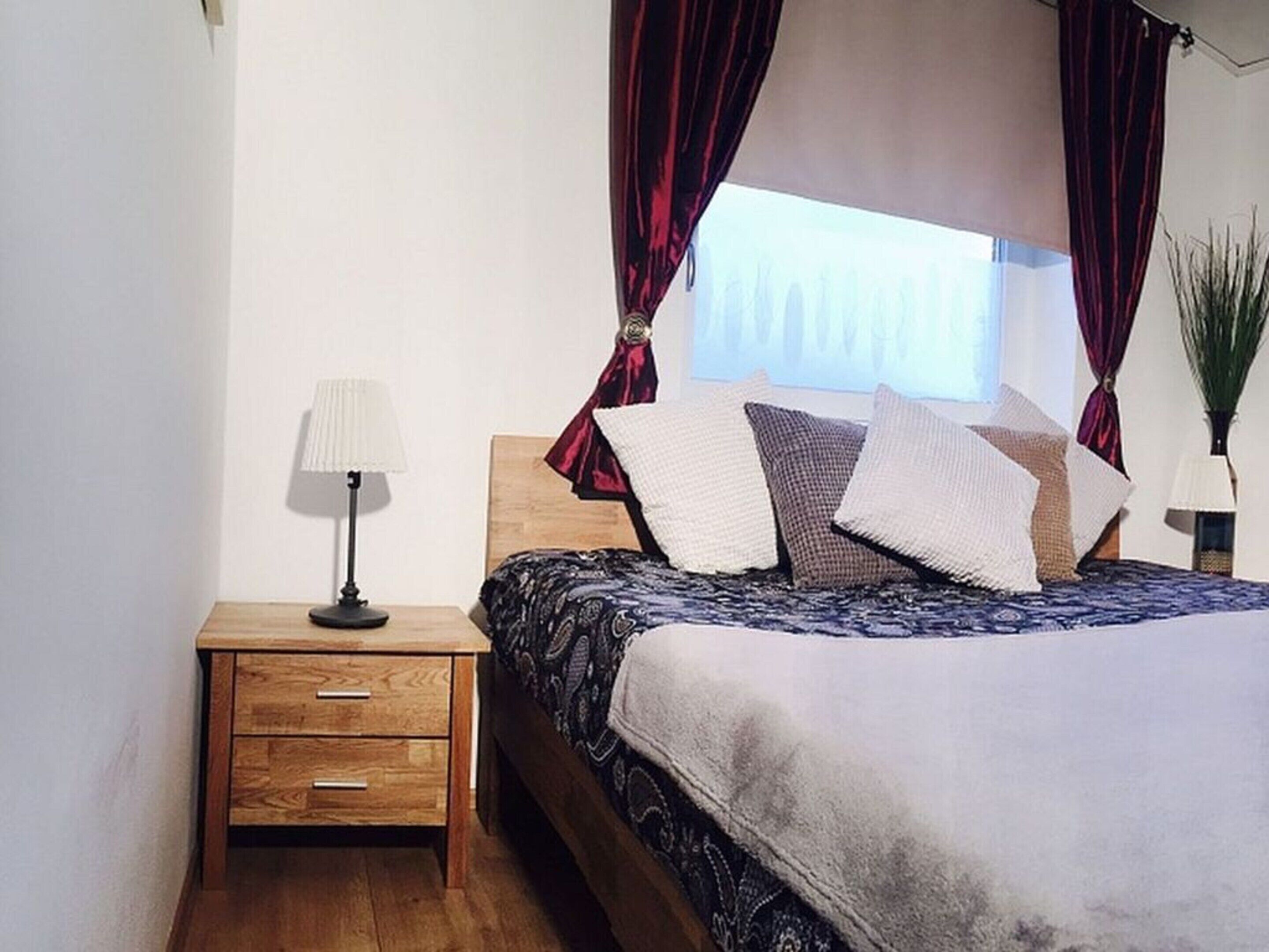 A bedroom with a wooden nightstand, a lamp, a bed with patterned bedding, and red curtains.