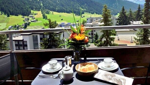 Una mesa lista para el desayuno, con vistas a la montaña, un florero con flores y una postal.