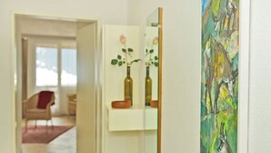Un pasillo con una pared blanca, un espejo que refleja un jarrón con una rosa y un cuadro.