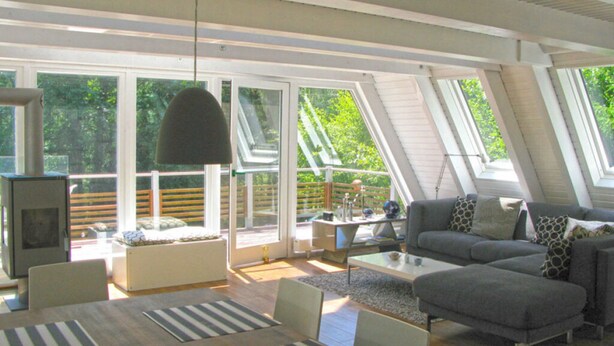 Een ruime woonkamer met een moderne bank, een eettafel en een open haard. Grote ramen zorgen voor veel natuurlijk licht.