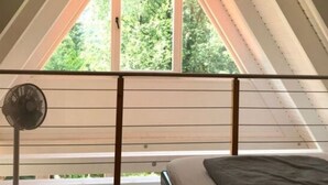 Een kamer met een schuine kap, een bed, een ventilator en een raam met uitzicht op groen.