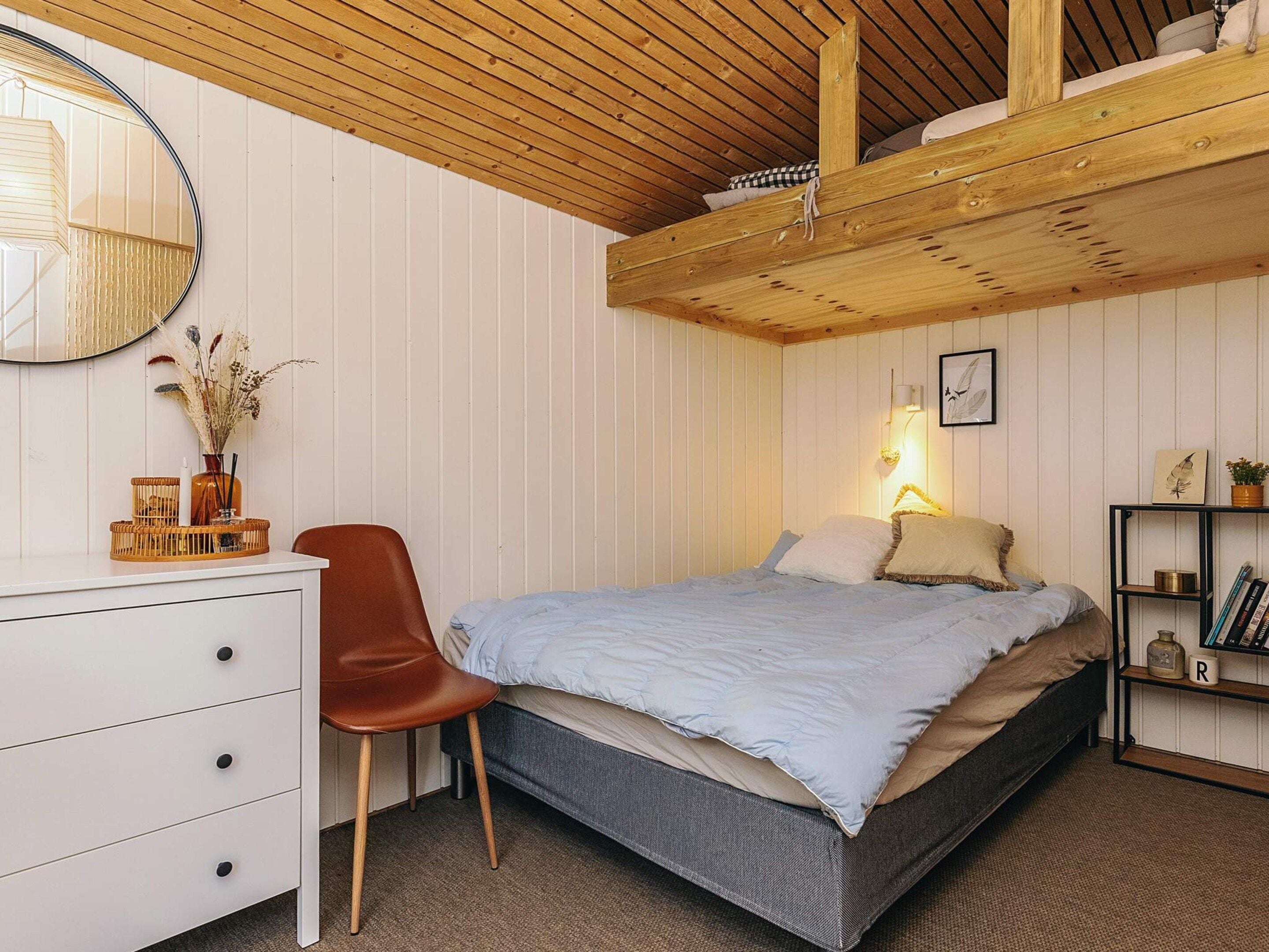 A bedroom with a bed, a desk, a chair, a mirror, a shelf with books, and a wall-mounted lamp.