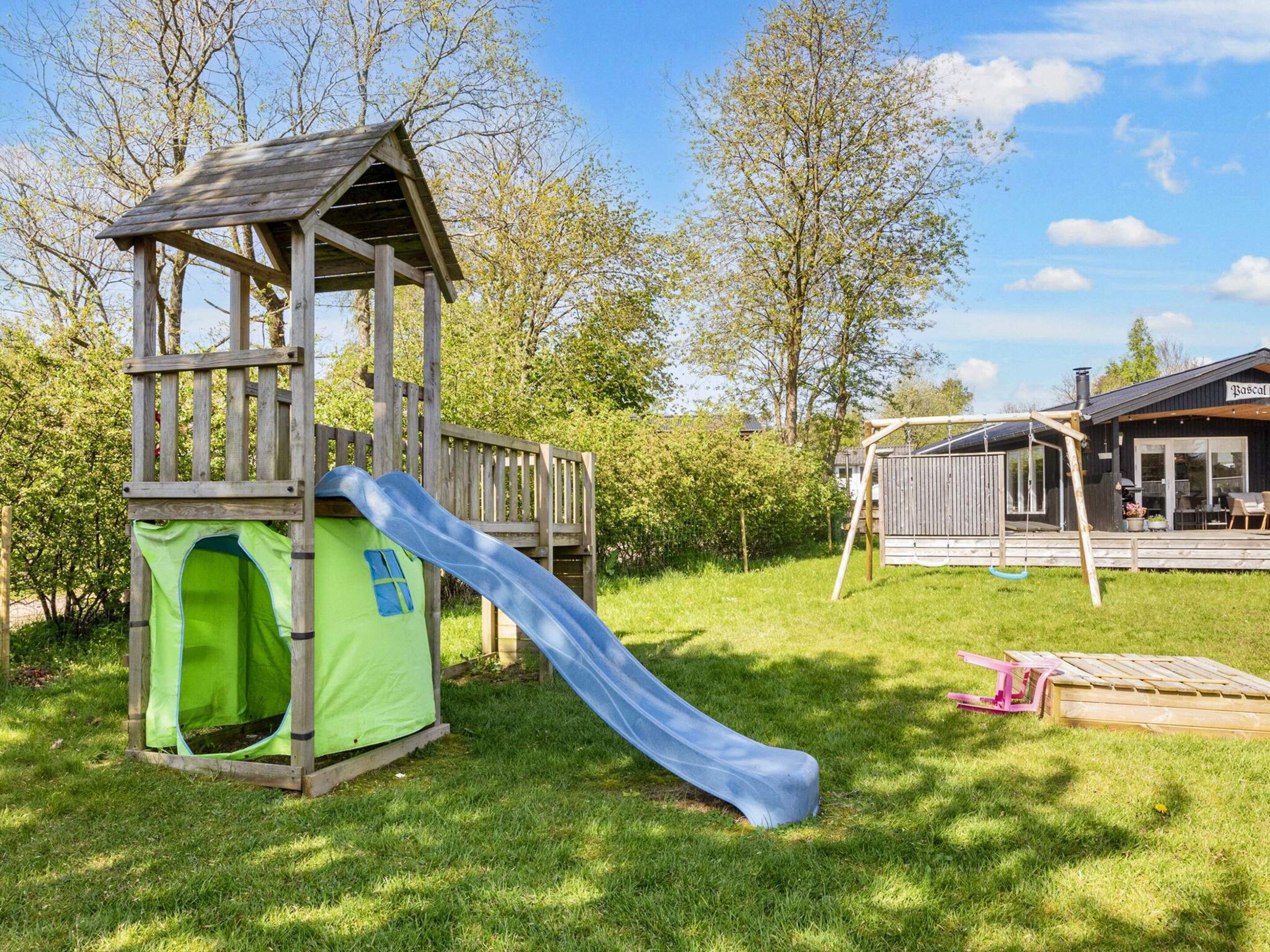 Une structure de jeu en bois, dotée d’un toboggan et d’une petite tente, installée dans un espace verdoyant avec des arbres et un bâtiment en arrière-plan.