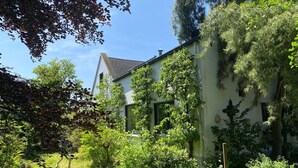 Une maison avec un toit en pente, entourée d’une végétation luxuriante et d’arbres.