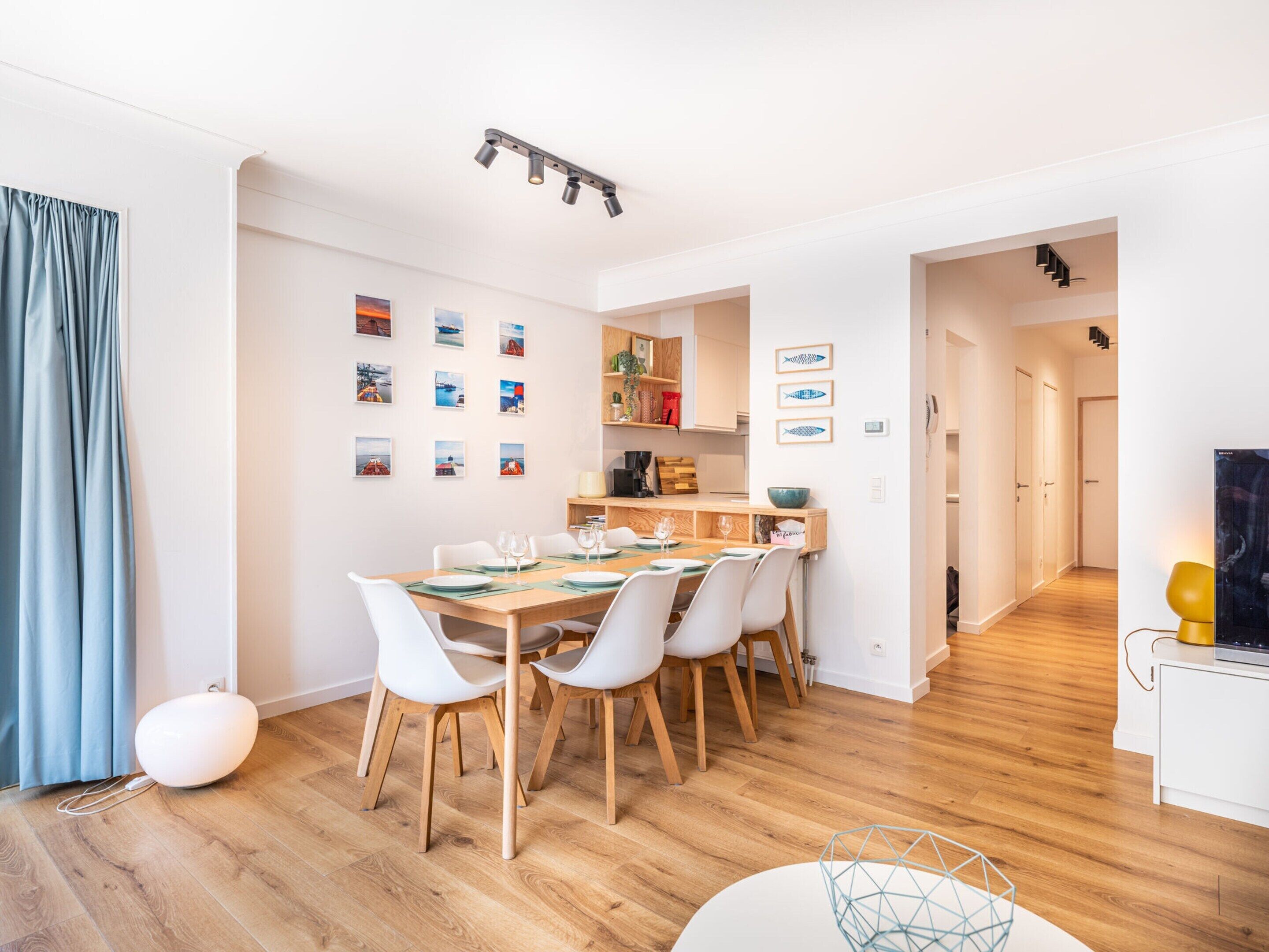 A modern dining area with a wooden table set for six, white chairs, and a kitchen in the background.