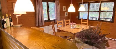 A wooden cabin interior with a dining table, chairs, a bar area with bottles, and hanging pendant lights.