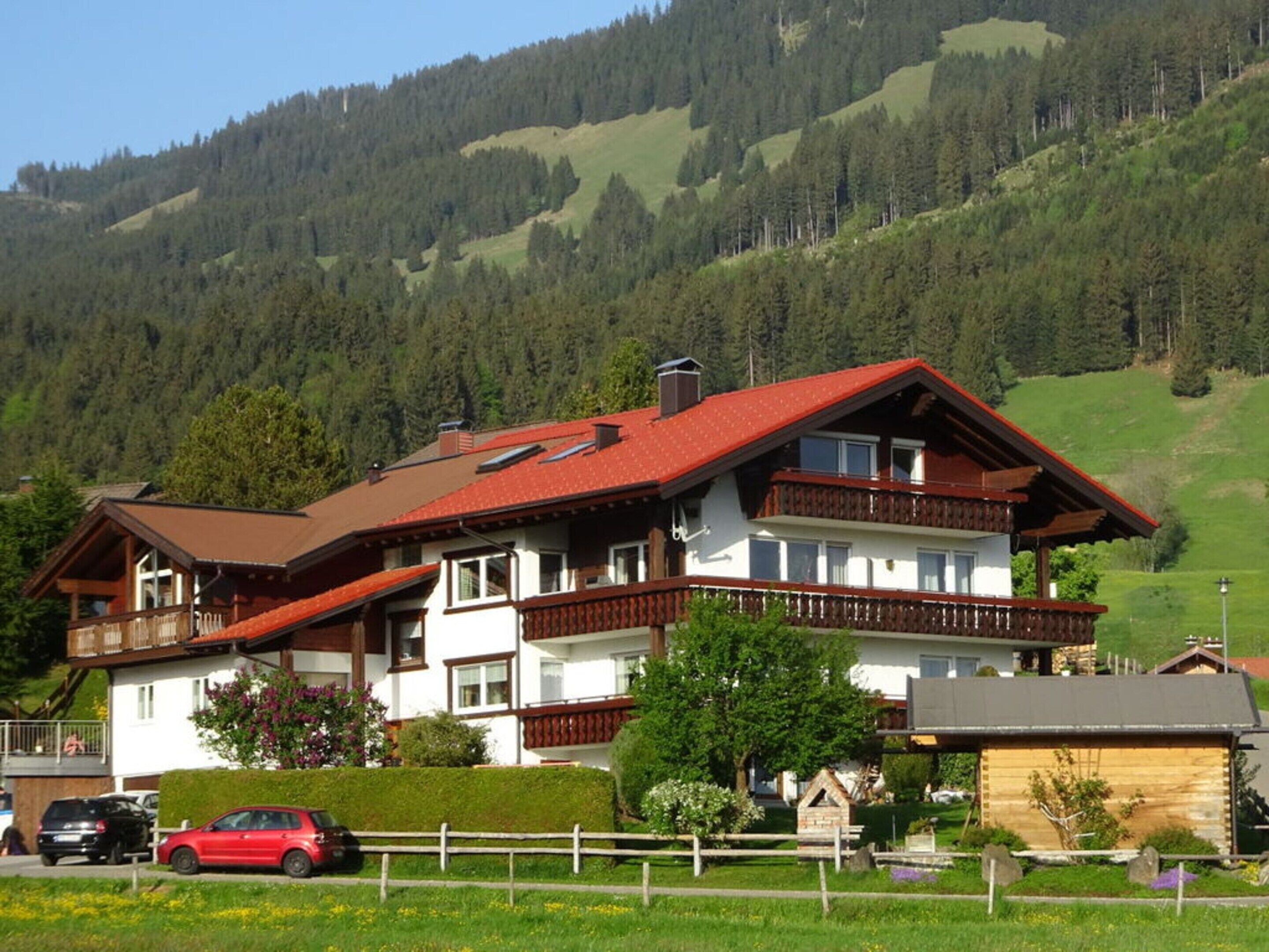 A two-story chalet with a red roof, wooden balconies, and a view of a forested mountain.