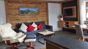 Un soggiorno moderno con una parete in legno, un divano con cuscini rossi e bianchi, una poltrona bianca e soffice, un tavolino e un televisore fissato alla parete.