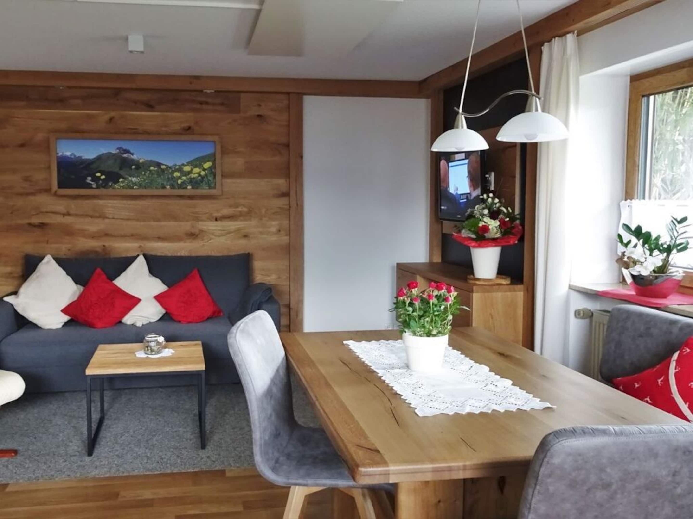 A cozy room with a wooden wall, a grey sofa with red and white cushions, a wooden dining table with a lace tablecloth, and a potted plant with red flowers.