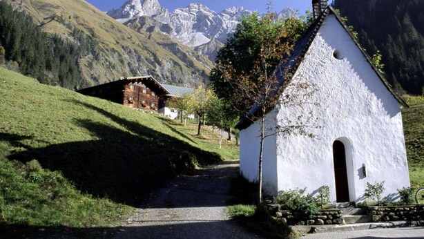 Una piccola chiesa bianca con un campanile, immersa in un paesaggio rurale con montagne sullo sfondo.