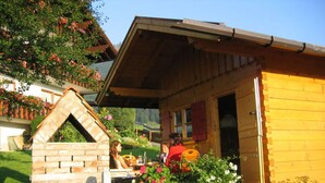 Una rustica baita di legno con un camino in pietra, immersa nel verde e fiorita.