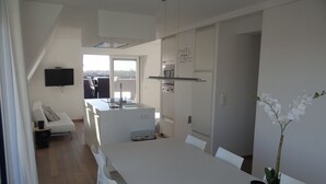 A modern kitchen with white cabinets, a built-in oven, and a microwave. There is a dining area with a white table and chairs, and a potted orchid on the table. - Apartment in Nieuwpoort With Terrace (Nieuwpoort)