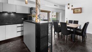 A modern kitchen with a central island, black countertops, and white cabinetry. There is a dining area with a table and chairs, and a large window providing natural light. - Villa in Istria With Heated Eco Pool (Brtonigla)