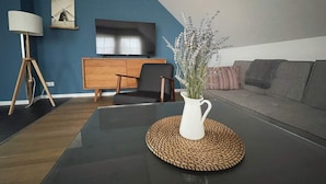 Un salon moderne avec une table basse en verre noir, un vase blanc orné de fleurs, un meuble en bois, une chaise noire, un canapé gris et un téléviseur à écran plat fixé au mur.