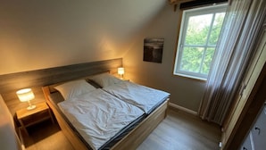 Une chambre à coucher avec un lit en bois, deux tables de chevet avec des lampes, une fenêtre avec des rideaux et une vue sur la verdure à l’extérieur.