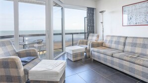 Una sala de estar moderna con un gran ventanal corredizo que da a un balcón con vista al mar. Hay dos sofás y una pequeña mesa con sillas.