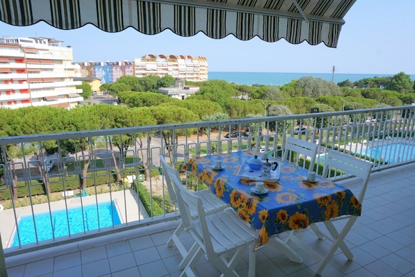 A balcony with a table set for two, overlooking a swimming pool and a view of buildings and the sea.
