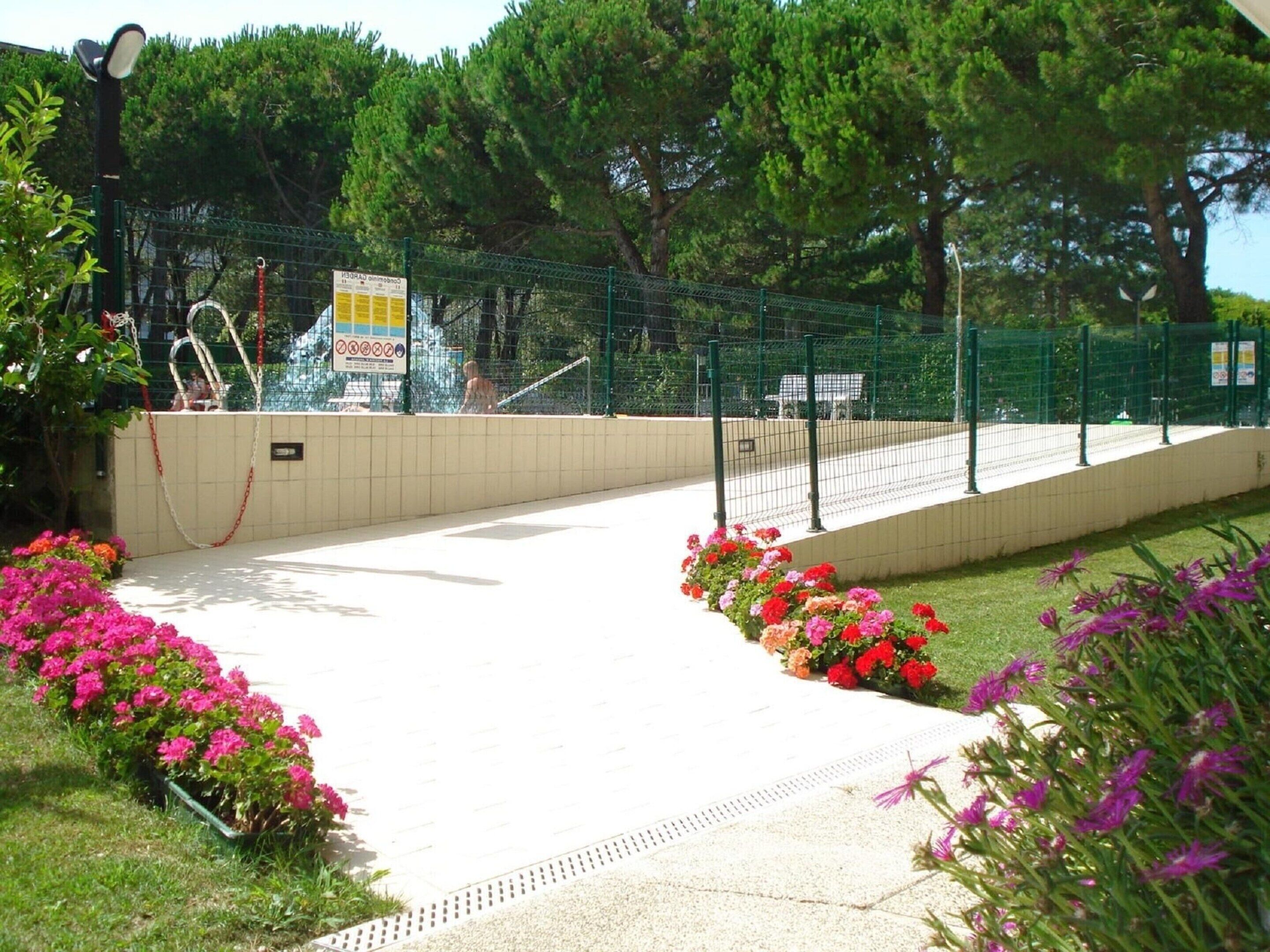 Un sendero que conduce a una zona cercada con un cartel, rodeada de plantas y árboles florecientes.