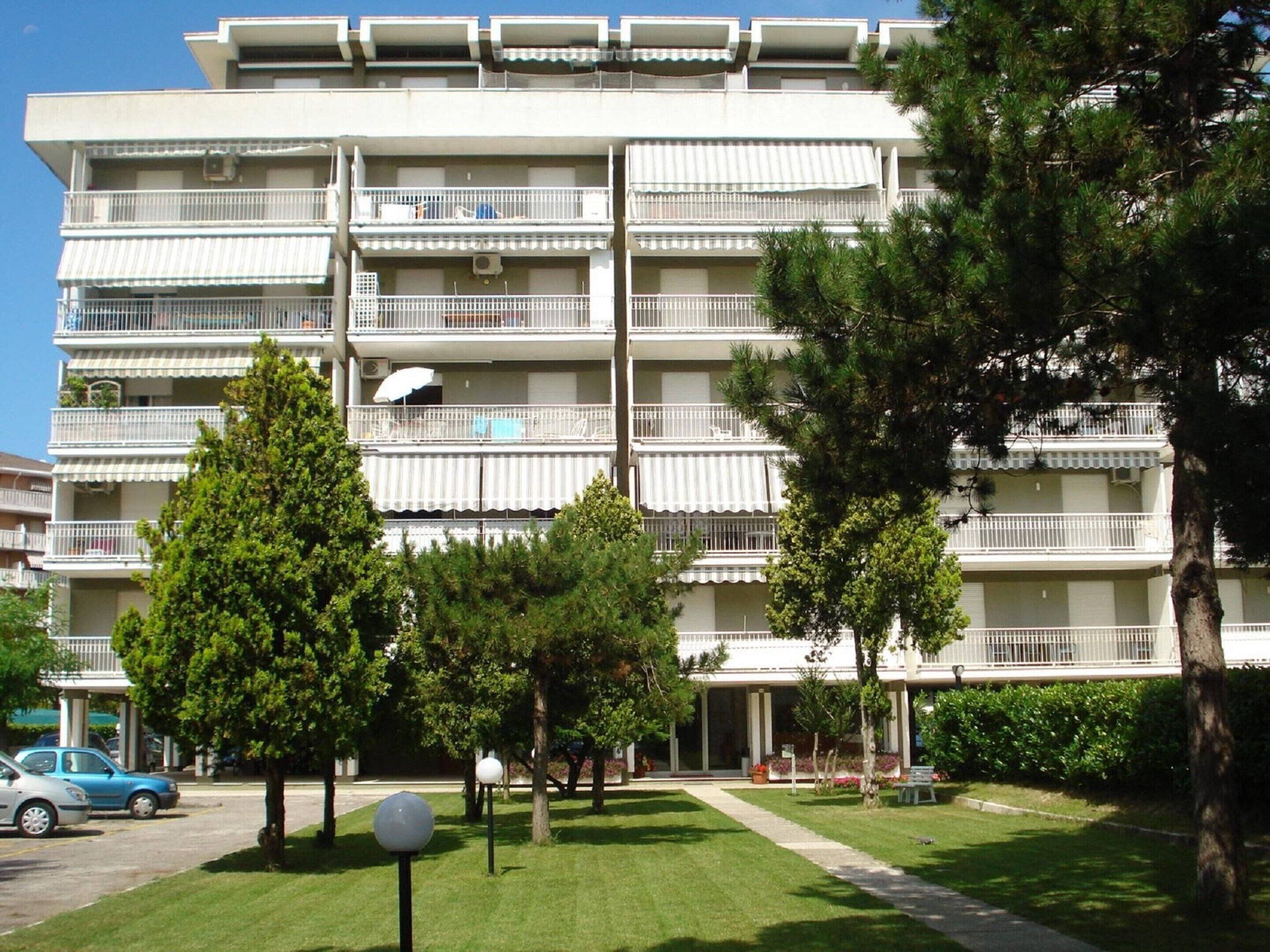 A multi-story residential building with balconies, a parking area, and a well-maintained lawn.