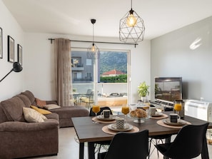 Un salon moderne avec un canapé, une table à manger et un téléviseur. Il y a un balcon d’où l’on peut admirer les bâtiments et la verdure environnante.