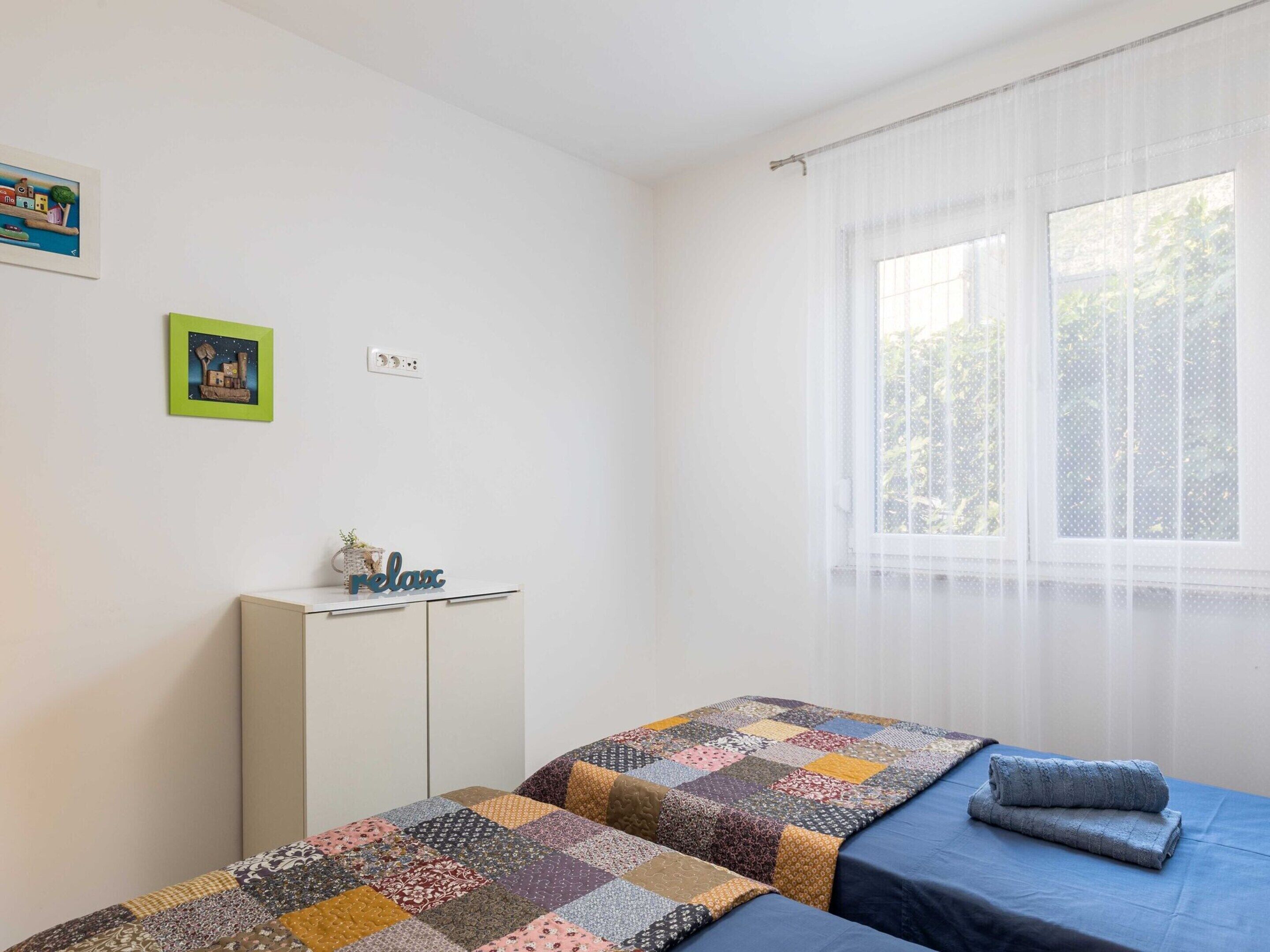 A bedroom with two beds, a small cabinet, and a window with sheer curtains.