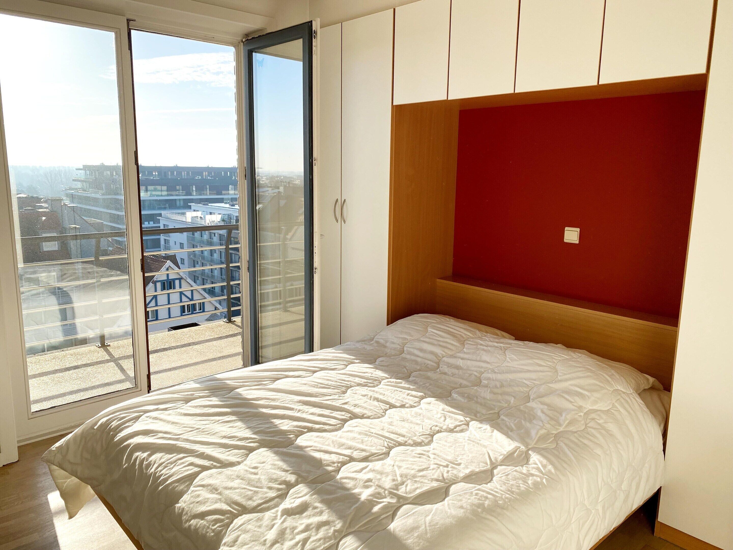 A bedroom with a bed, a red accent wall, and a view of buildings through a sliding door.