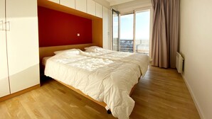Un dormitorio con una cama grande, suelo de madera, una pared de acento roja y vistas a la ciudad a través de amplias ventanas.