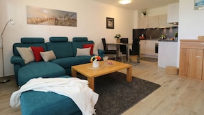 A modern living room with a teal sofa, a wooden coffee table, and a dining area with a small kitchen in the background.