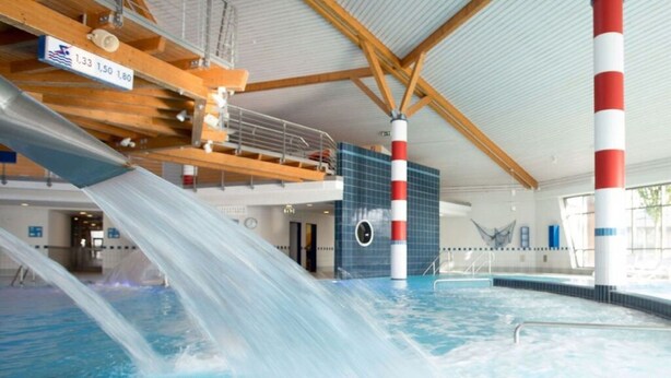 Una piscina interior con tobogán de agua, vigas de madera y pared de azulejos azules.