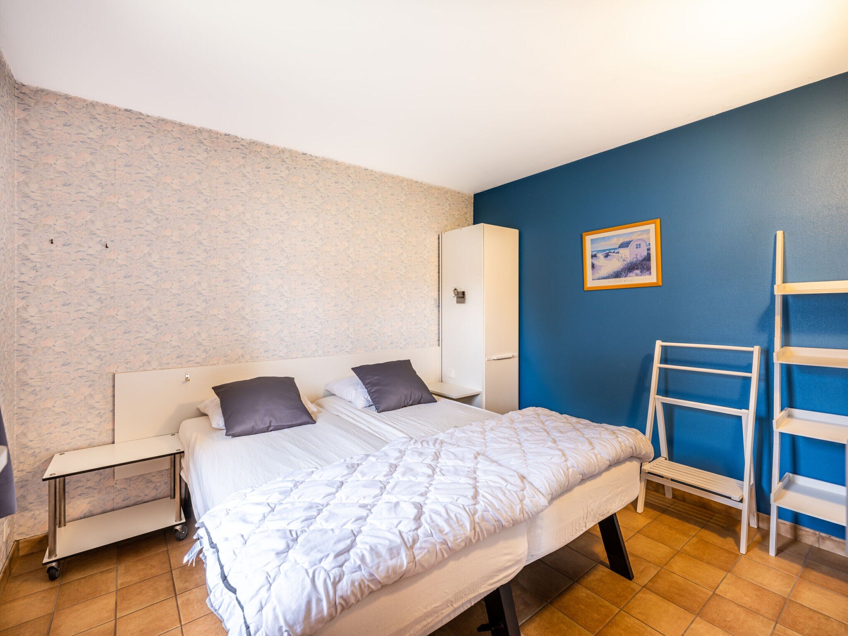 Un dormitorio con cama, mesita de noche, armario, escalera y un cuadro en la pared.