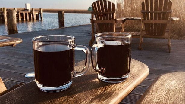 Two glass mugs on a wooden table by a waterfront with chairs and a dock in the background. - Holiday Apartment Dune Dream (Zingst)