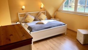 A bedroom with a sloped ceiling, a bed with white headboard, two bedside lamps, a window with a view of greenery, and a wooden table. - Holiday Apartment Dune Dream (Zingst)