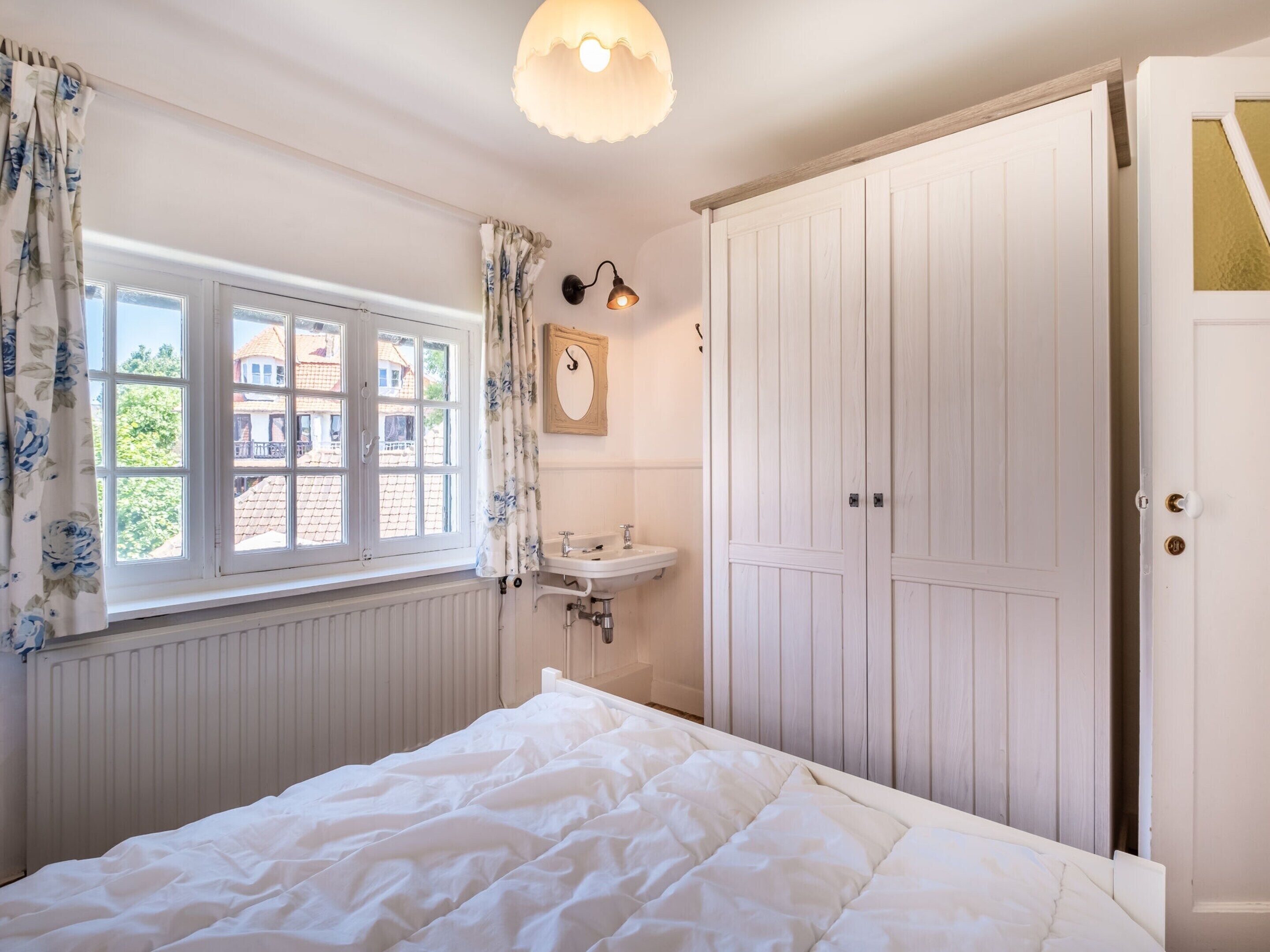 A bedroom with a bed, a window with curtains, a bedside table, a mirror, and a wardrobe.