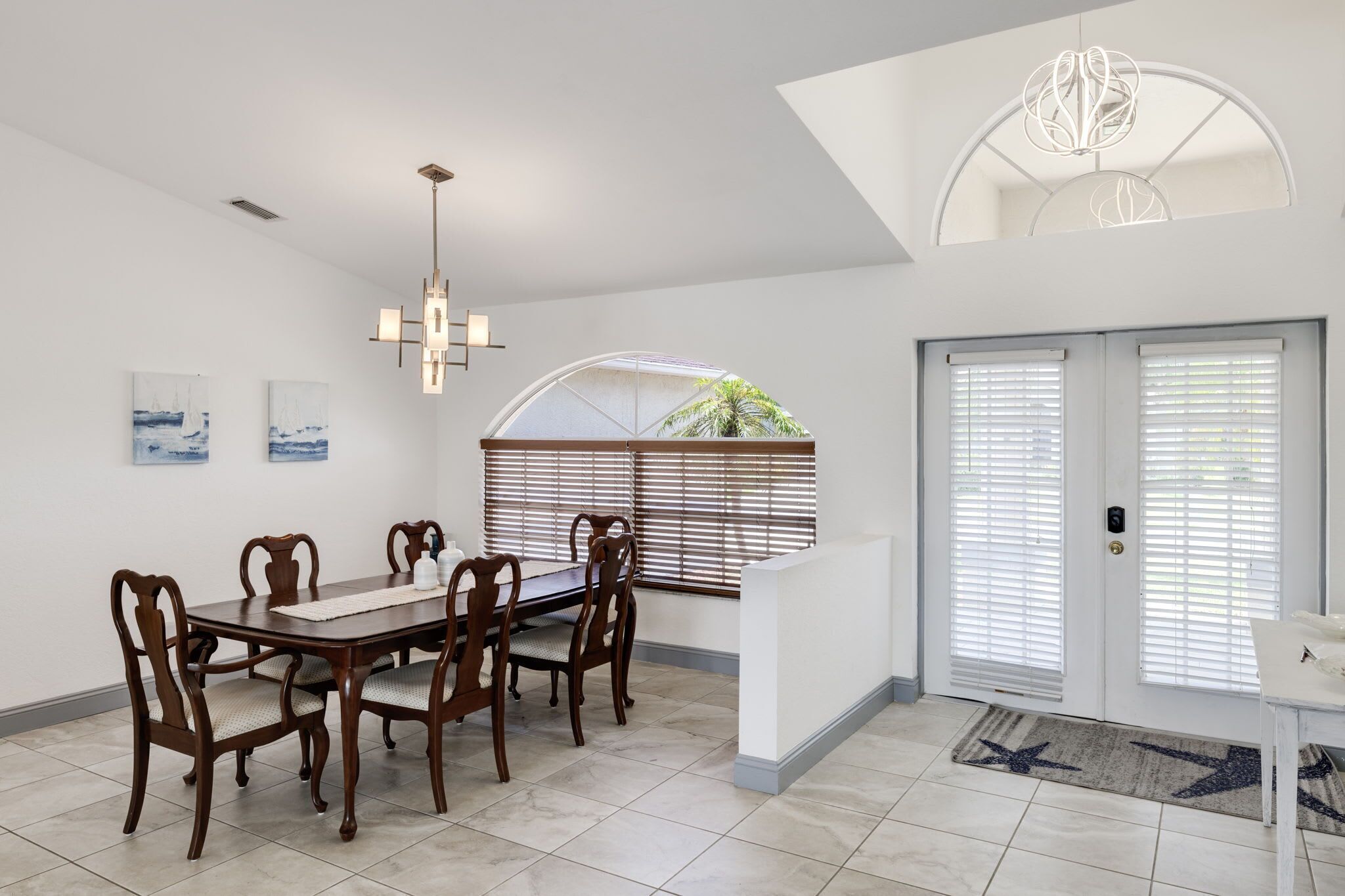 A dining area with a wooden table and chairs, a modern chandelier, and a view through a window with blinds.