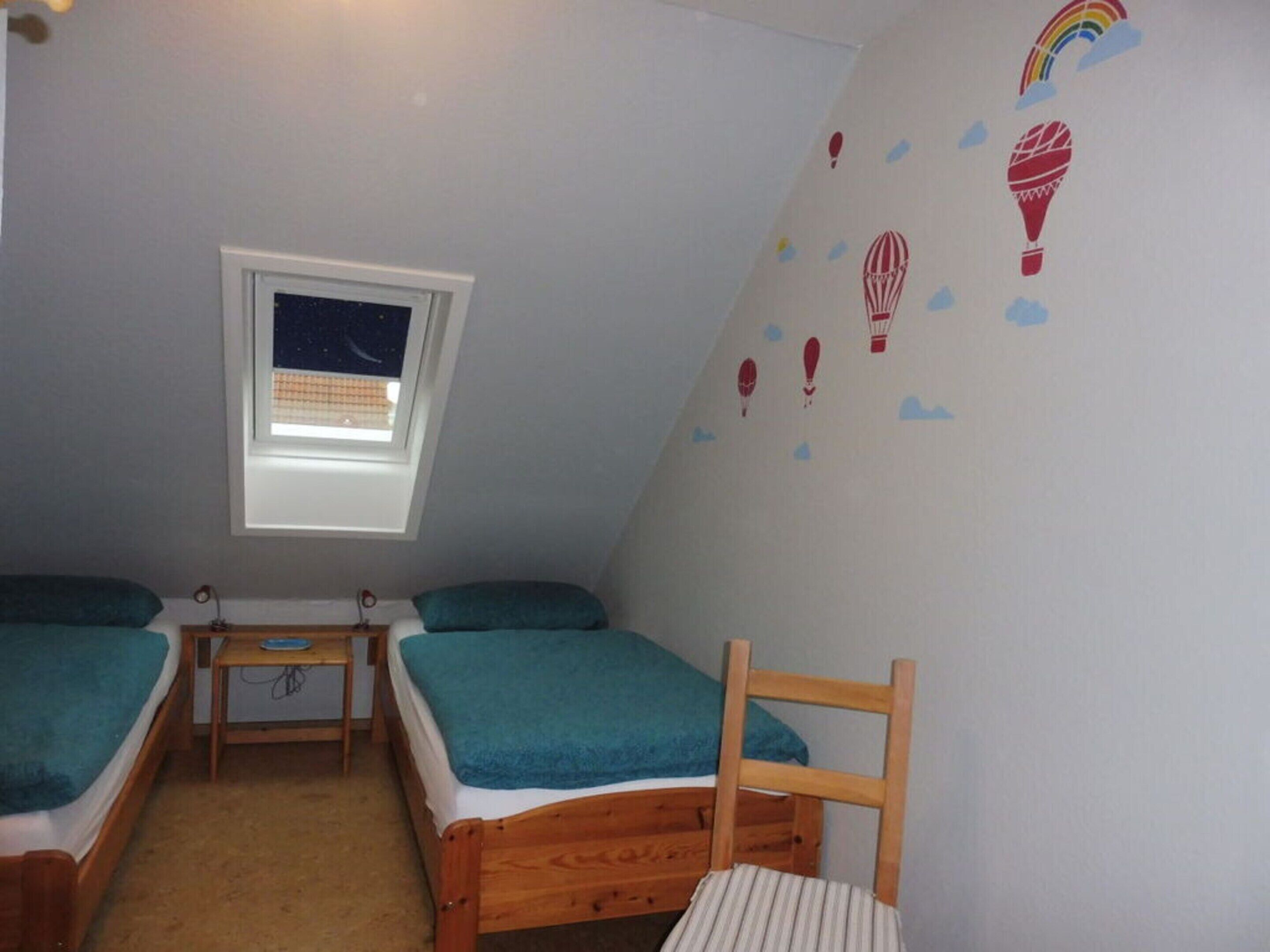 Ein kleiner, gemütlicher Raum mit zwei Einzelbetten, einem Holzsessel, einem kleinen Tisch und einem Fenster mit Blick auf den Nachthimmel.