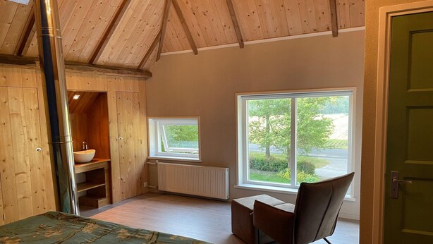 A room with a wooden ceiling, a large window with a view of greenery, a bed with a floral bedspread, and a chair.