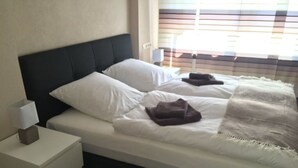A bedroom with a bed, white nightstands, and a window with blinds. - Holiday Apartment Wurzburg (Würzburg)
