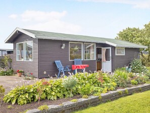 A small, single-story house with a dark exterior, a front porch with two blue chairs, and a red table. - 5 Person Holiday Home in Saeby (Sæby)