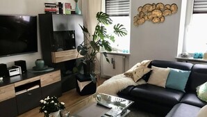 A modern living room with a flat-screen TV, a glass coffee table, a black sofa with decorative pillows, a potted plant, and a bookshelf. - Holiday Apartment Gertie (Saarburg)