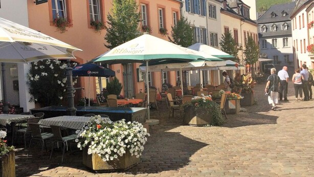A cobblestone street with outdoor seating and umbrellas, surrounded by traditional European buildings. - Holiday Apartment Gertie (Saarburg)