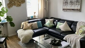 A modern living room with a black leather sofa, a glass coffee table, and a large window with curtains. - Holiday Apartment Gertie (Saarburg)