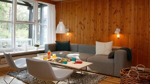 A cozy living room with a wooden wall, a grey sofa, a white armchair, a small table with snacks, and a wicker basket. - 6 Person Holiday Home in Vaeggerlose (Vaeggerlose)