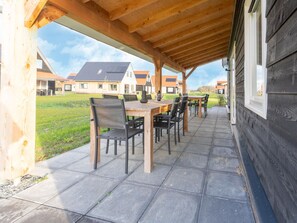 Um pátio externo com mesa e cadeiras de madeira, uma pérgola acima e vista para edifícios residenciais ao fundo.