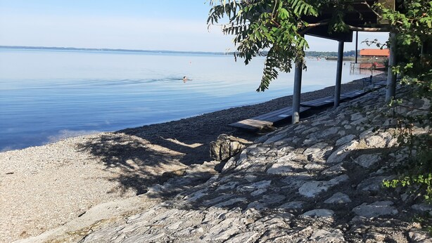 A rocky shoreline with a calm body of water, a wooden deck, and a building with a red roof in the background.