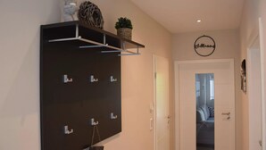 A hallway with a dark wall-mounted board, a small shelf with decorative items, and a door leading to a bedroom. - Beach Apartment in Schleswig-holstein (Westerdeichstrich)