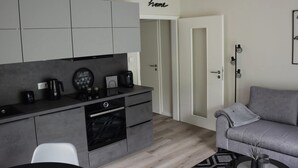 A modern kitchen with grey cabinets, a coffee machine, and a built-in oven. There is a grey sofa and a small table with a plant. - Beach Apartment in Schleswig-holstein (Westerdeichstrich)